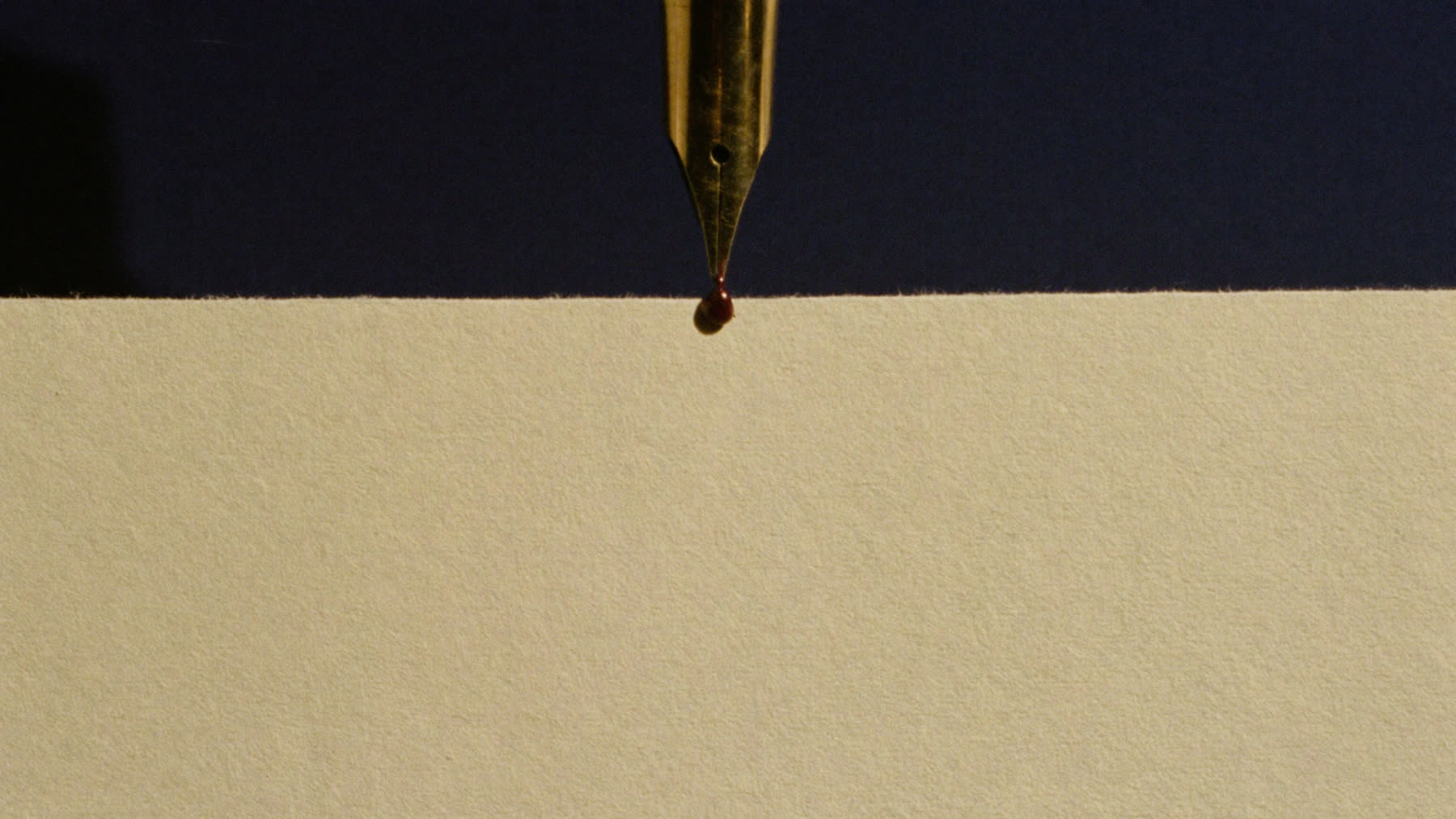Macro photograph of a gold fountain pen nib with a bead of oxblood ink over cream paper